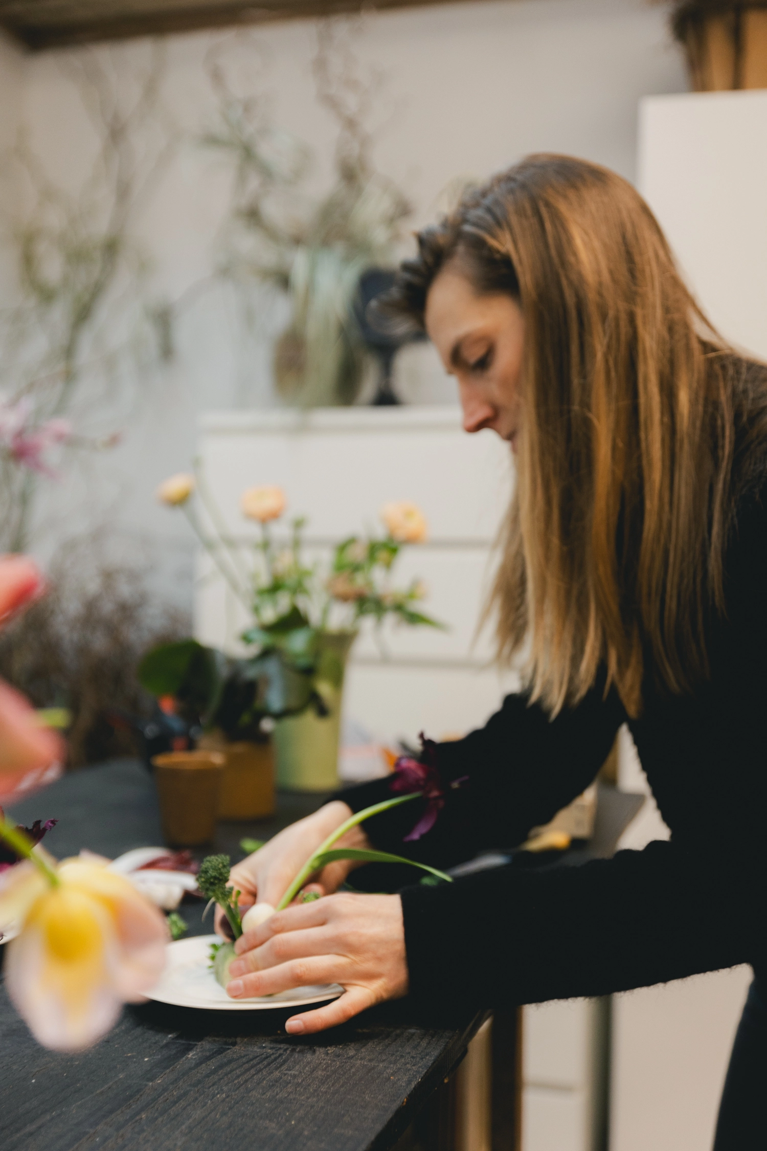 Une femme se tenant début face à un plan de travail, elle compose avec des fleurs et des légumes pour agrémenter une table.