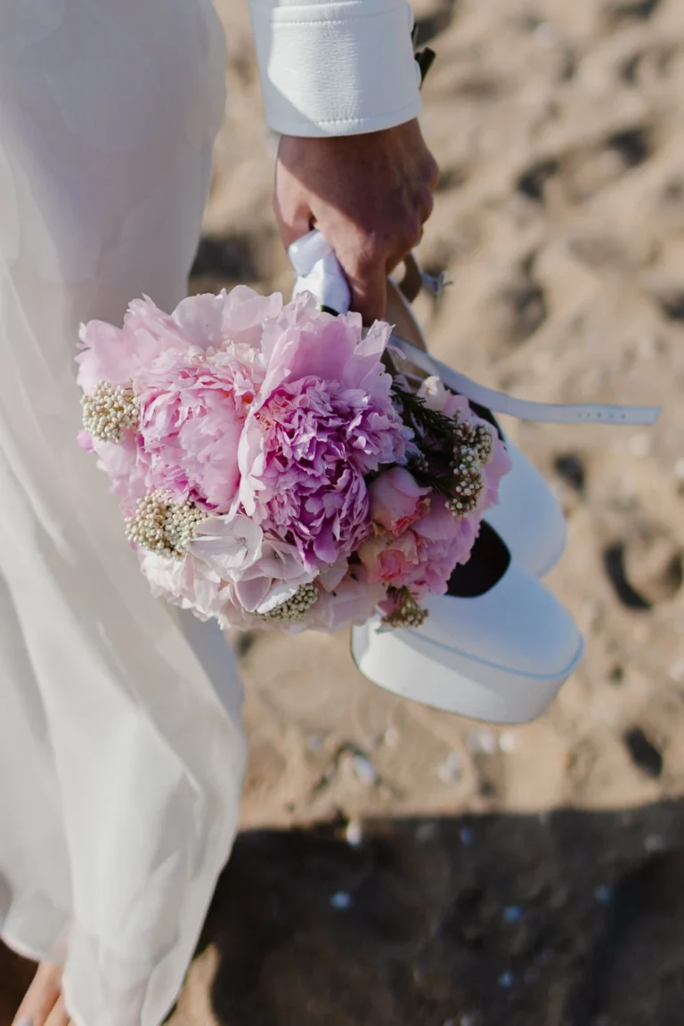 Un main tient un bouquet de fleur et ses chaussures pour marcher dans le sable chaud.