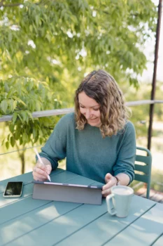 Une femme est assise en extérieur, elle est penché sur sa tablette graphique et tenant dans sa main un stylet.