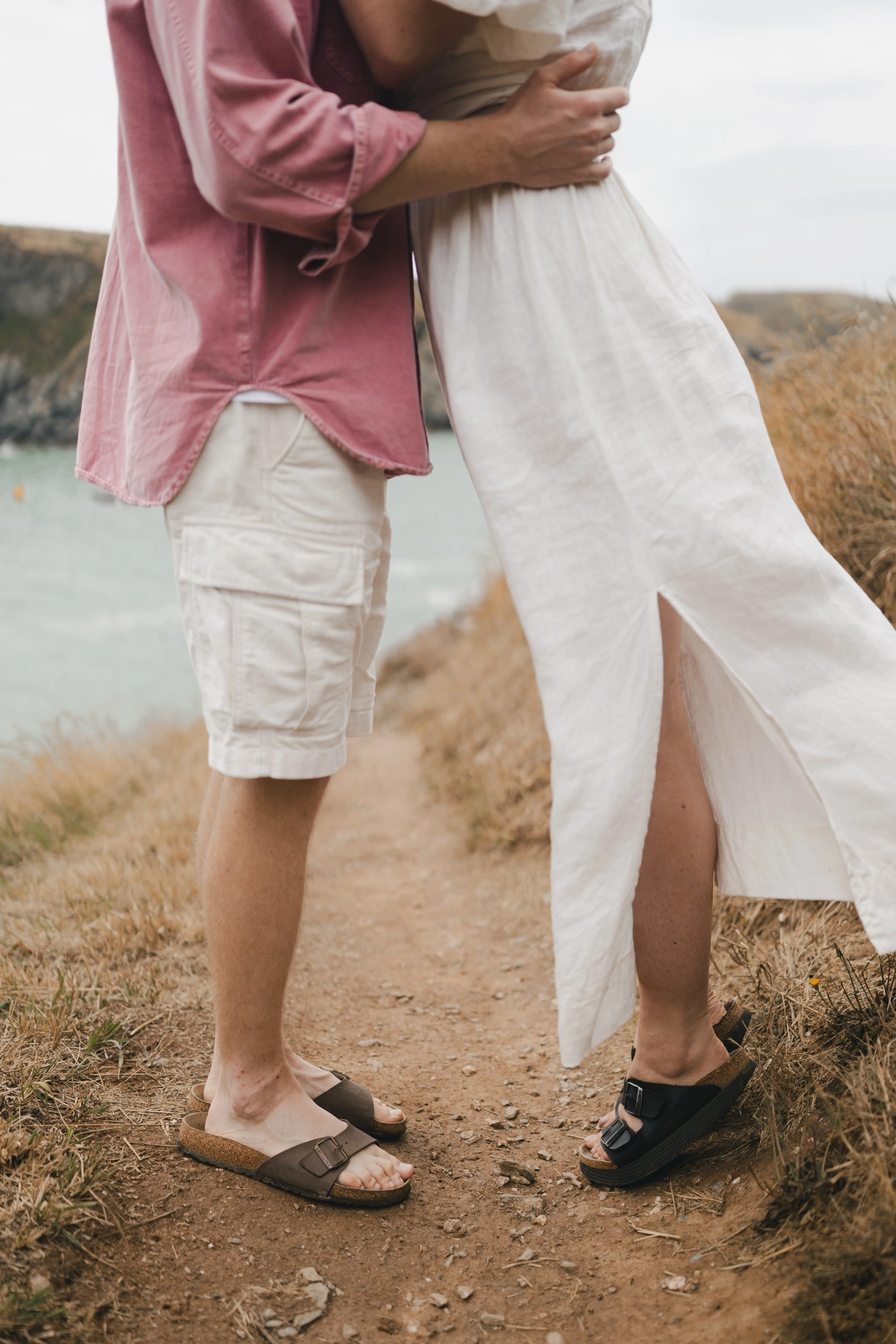 Le long d'un chemin, un homme et une femme portant une robe fluide - où le vent s'immisce, se tenant l'un en face de l'autre, il tient à la taille sa compagne et elle se tient sur la pointe des pieds.