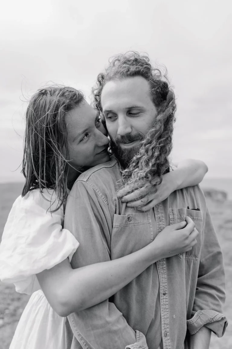 Une femme porte ses bras par derrière un homme sur son torse, le regarde et lui est légèrement tourné pour voir sa compagne.