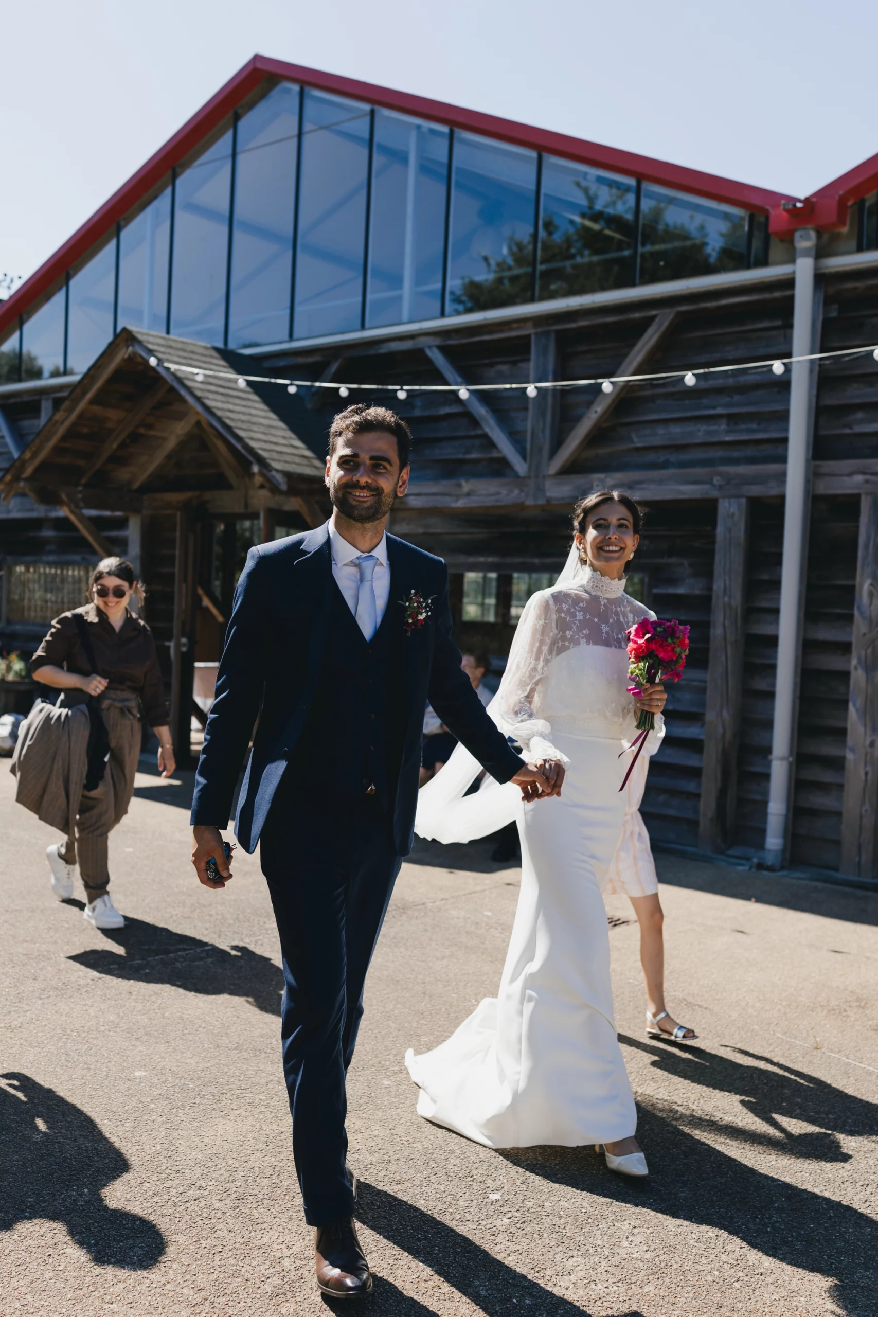 Une couple marche main dans la main, elle est vêtue d'une robe blanche et lui en costume. En arrière-plan on aperçoit les Serres de Rospico.