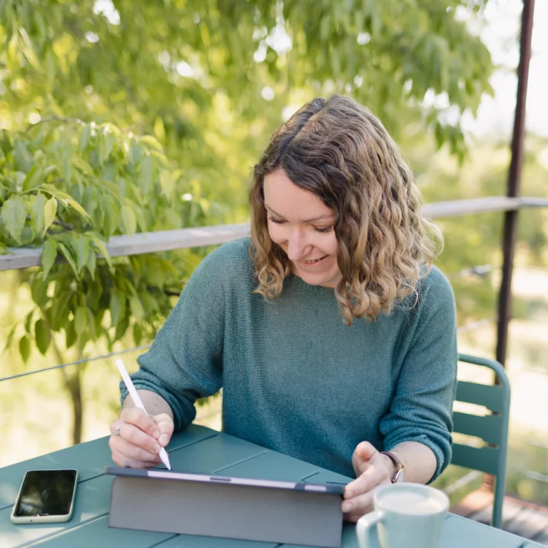 Une femme est assise en extérieur, elle est penché sur sa tablette graphique et tenant dans sa main un stylet.