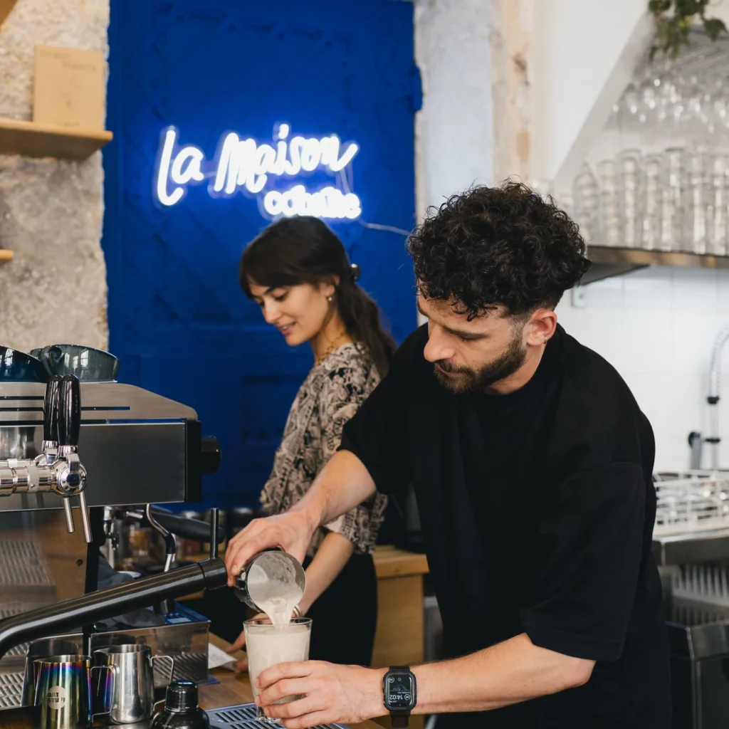 Valentin au premier plan qui créer un Chaï Latte et Mélanie au second plan qui prépare une commande.